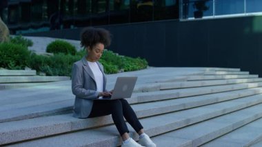 İş kadını modern taş basamaklarda oturuyor, kentsel bir ortamda dizüstü bilgisayara odaklanıyor, uzaktan çalışmayı ve üretkenliği simgeliyor..
