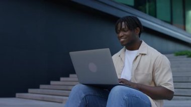 Genç adam dışarıda merdivenlerde dizüstü bilgisayar kullanarak oturuyor, rahat bir şehir ortamında çalışırken ya da çalışırken mutlu bir şekilde gülümsüyor..