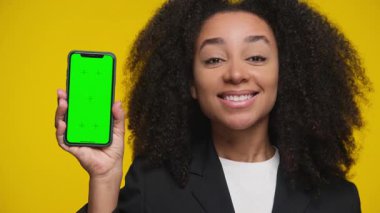 Kendine güvenen bir kadın, yeşil ekranlı bir akıllı telefonu sarı arka planda tutarken gülümseyip onaylayarak başını sallıyor..