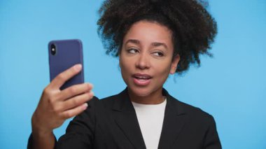 Genç bir kadın video görüşmesi yapıyor, elinde akıllı telefon var ve mavi arka plandaki ekrana bakarken konuşuyor..