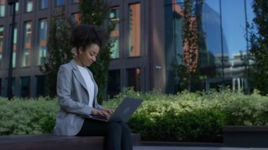 Modern bir açık hava çalışma sahasında bir bankta otururken dizüstü bilgisayarı üzerinde çalışan iş elbiseli genç bir kadın..