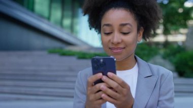 Spor ceketli genç bir kadın dışarıda akıllı telefonuyla yazarken gülümsüyor, modern basamakların ve binaların yanında duruyor..