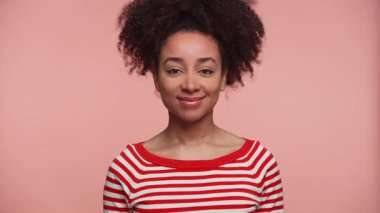 Kendine güvenen kıvırcık saçlı, kameraya hafifçe gülümseyen, pembe arka planda kırmızı ve beyaz çizgili gömlek giyen genç bir kadın..