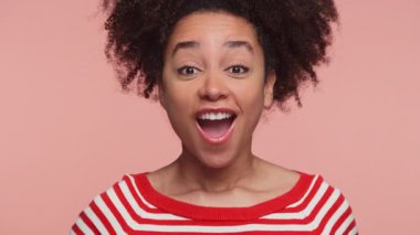 Çizgili tişörtlü neşeli genç kadın heyecan içinde yumruğunu kaldırıyor, başarısını, kutlamasını ya da pembe arka plana karşı enerjik pozitifliğini ifade ediyor..