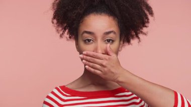 Tek eliyle ağzını kapatan çizgili tişörtlü, şaşırmış genç bir kadın, pembe arka planda izole edilmiş şok, gizlilik veya suskunluk belirtileri gösteriyor.