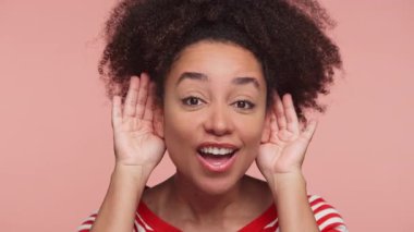 Gülümseyen genç bir kadın, ellerini kulaklarına dayayıp, sanki pembe arka planda izole edilmiş, net bir şey duymaya çalışıyormuş gibi ilgi ve ilgi gösteriyor.