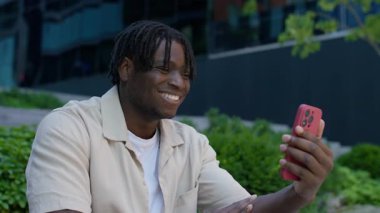 Mutlu genç adam dışarıda video görüşmesinden zevk alıyor, elinde akıllı telefon ve modern bir ofis binasının önünde gülümsüyor..