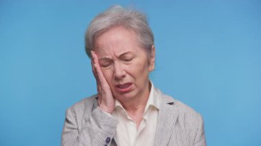 Gri saçlı ve çizgili ceketli yaşlı bir kadın. Yüzünü rahatsızlık içinde tutuyor. Mavi arka planda acı ya da duygusal sıkıntı gösteriyor..