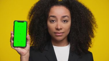 Kendine güvenen iş elbiseli bir kadın yeşil ekranlı akıllı bir telefon gösteriyor. Uygulama modelleri veya ürün yerleştirme için mükemmel, sarı arka planda izole edilmiş..