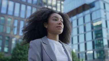 Spor ceketli genç bir kadın modern şehir bölgesinde yürüyor, düşünceli ve cam ofis binalarının yanında kararlı görünüyor..