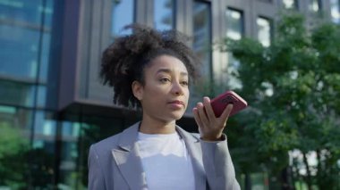 Spor ceketli genç bir kadın dışarıda akıllı telefonuna konuşuyor, muhtemelen ses asistanı kullanıyor, arkasında modern binalar var..