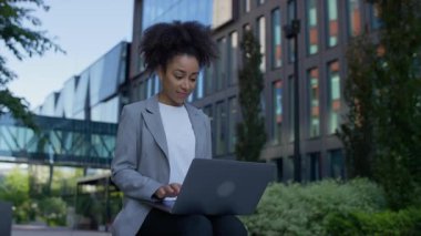 Modern bir ofis kampüsünde dizüstü bilgisayar kullanan genç profesyonel bir kadın yeşillik ve binalarla gözler önüne serildi..