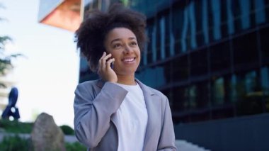 Spor ceketli neşeli genç bir kadın arka planda cam binalar olan modern bir şehir ortamında dışarıda telefonla konuşuyor..