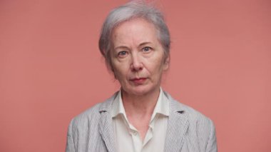 Gri saçlı, tarafsız bir ifade takınan, pembe arka planda duran, çizgili ceket ve beyaz gömlek giyen yaşlı bir kadın..