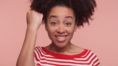 Kırmızı çizgili tişörtlü neşeli genç bir kadın geniş gülümsüyor ve heyecan içinde yumruğunu kaldırıyor. Pembe arka plandaki zaferi kutluyor..