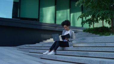 Profesyonel bir genç kadın dışarıda merdivenlerde dizüstü bilgisayarla oturmuş, modern bir ofis binasının yakınındaki bir defterdeki notları inceliyor..