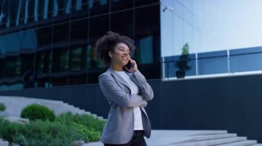 Gri ceketli genç bir kadın telefonla konuşurken gülümsüyor, modern cam ofis binasının önünde duruyor..