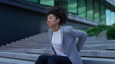 İş kadını dışarıda merdivenlerde oturuyor. Rahatsızlık içinde belini tutuyor. Acı ve fiziksel gerginlik belirtileri gösteriyor..