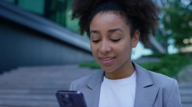 Spor ceketli genç bir kadının, şehir ortamının dışında beklerken, akıllı telefonuyla mutlu bir şekilde kayışının yakın çekimi..