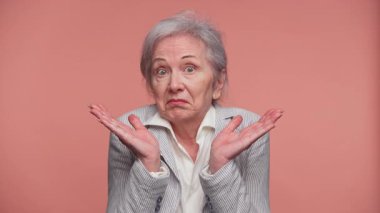 Çizgili ceketli yaşlı bir kadın ellerini kaldırarak omuz silkiyor ve şaşkın bir yüz ifadesi takınıyor, düz pembe bir zemine karşı duruyor..