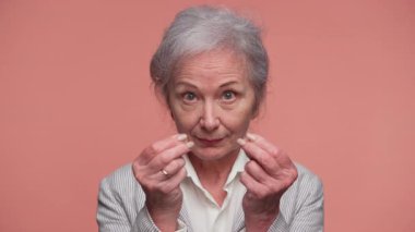 Çizgili ceketli yaşlı bir kadın parmak masajı yapıp pembe arka planda kameraya bakıyor..