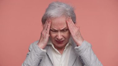 Çizgili ceketli yaşlı bir kadın başını acı ve hüsran içinde tutuyor. Pembe arka planda baş ağrısı belirtileri gösteriyor..