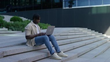 Modern bir binanın dışındaki beton basamaklarda oturan genç bir adam, rahat bir şehir ortamında dizüstü bilgisayarıyla çalışmaya odaklanıyor..