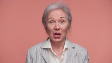 Neşeli yaşlı bir kadın heyecanla tepki veriyor, geniş bir gülümseme ve pembe arka planda izole edilmiş iyi haberler aldıktan sonra kutlamada yumruğunu kaldırıyor..