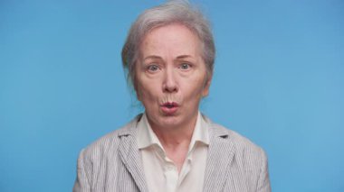 Olumsuz şok ya da inanmazlık gösteren yaşlı bir kadının portresi, açık ağızlı ve mavi arka planda geniş gözlü..