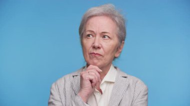 Düşünceli bir yüz ifadesi ve çenesi üzerinde eli olan yaşlı bir kadın, mavi arka planda derin derin düşünürken ya da düşünürken yana bakıyor..