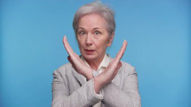 Üst düzey bir kadın, reddedilme, onaylama ya da durmama sinyali vermek için kollarını X ile kavuşturuyor. Mavi arka planda sert bir ifadeyle..