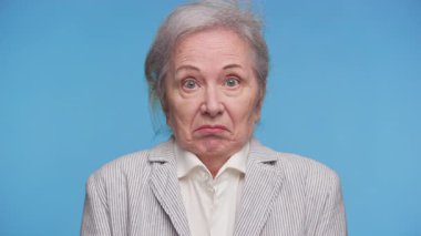 Omuzları kalkık ve kafası karışmış yaşlı bir kadın, düz mavi bir arka plana karşı kafa karışıklığı ya da kayıtsızlık taşıyor..