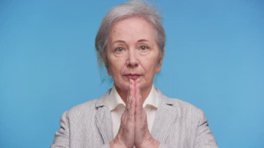 Kıdemli kadın yüzün önünde el ele tutuşuyor, dua ediyor, saygı gösteriyor, ya da saf mavi bir arka plana kafa yoruyor..