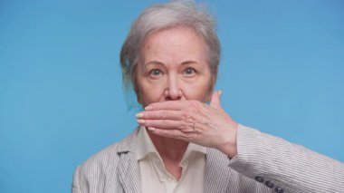 Kıdemli kadın elini ağzının üstünde tutuyor, şok geçiriyor, gizlilik veya pişmanlık duyuyor, sağlam bir mavi arka plana karşı..