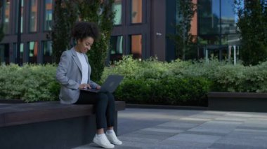 Kendine güveni tam genç bir kadın, güneşli bir günde modern bir ofis binasının önündeki bankta otururken internette gezinmeyi seviyor..