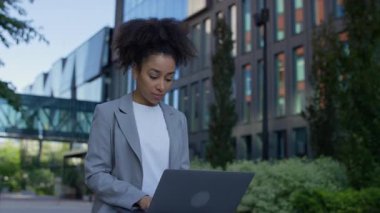 Ciddi bir iş kadını dışarıda modern cam ofis binasının yanında oturuyor, yeşil bir şehirde uzaktan çalışırken bilgisayarına konsantre oluyor..