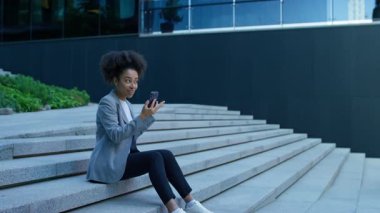 Genç, profesyonel bir kadın, elinde akıllı bir telefonla açık hava merdivenlerinde oturuyor ve modern bir ofis binasının yakınındaki bir video görüşmesine katılıyor..