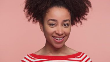 Kırmızı beyaz çizgili süveter giyen neşeli genç bir kadın pembe arka planda büyük bir gülümsemeyle kameraya göz kırpıyor..