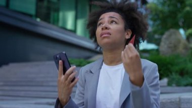 Çevrimiçi ödemelerde başarısız olduğu için üzgün görünen genç bir kadın, elinde akıllı telefon ve kredi kartı ile modern binanın yakınlarında hayal kırıklığı yaşıyor..