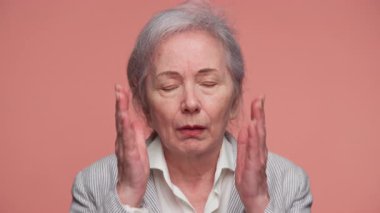 Gri saçlı, sıkıntılı yaşlı bir kadın. Şok olmuş ve endişeli görünüyor. Pembe bir arka planda yüz yüze..