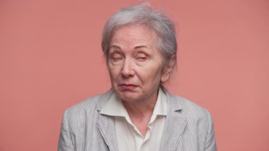 Gri saçlı yaşlı bir kadın onaylamadığını ya da anlaşmazlık yaşadığını söylüyor. Pembe arka plana ciddi bir bakışla başını hafifçe sallıyor..
