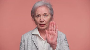 Yaşlı bir kadın açık avuçlarını kaldırıyor ya da hareketlerini durduruyor. Kararlılığını ya da pembe arka plana karşı tavır sergiliyor..
