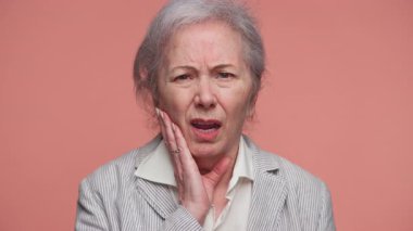 Çenesini tutarak acı çeken yaşlı bir kadın pembe arka planda diş rahatsızlığı ya da diş ağrısı belirtileri gösteriyor..