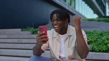 Mutlu genç adam dışarıda merdivenlerde oturuyor, gülümsüyor ve akıllı telefonundan harika haberler aldıktan sonra kutlamada yumruğunu kaldırıyor..