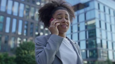 Modern cam ofis binalarının yakınında telefonla konuşurken gülümseyen gri ceketli genç profesyonel bir kadın..