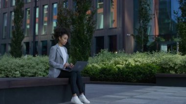 Açık havada dizüstü bilgisayarla çalışan gri ceketli genç bir kadın, şehir merkezinde bir bankta otururken düşünceli bir şekilde duruyor..