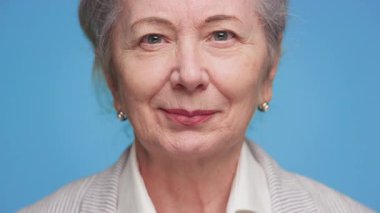 Kaşlarını kaldırmış, kurnaz bir gülüşü olan, mavi arka planda kamerayla doğrudan göz teması kuran yaşlı bir kadının yakın çekimi..