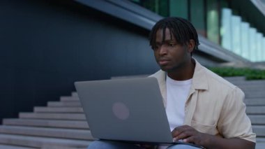 Genç adam modern bir binanın dışında merdivenlerde oturuyor, dizüstü bilgisayarı ile birlikte kentsel çalışma ortamında kayıyor..