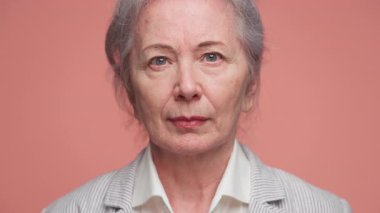 Gri saçlı, spor ceket giyen, pembe arka planda özgüven ve bilgelik gösteren yaşlı bir kadının yakın plan portresi..