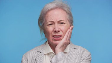 Yaşlı bir kadın yanağını rahatsızlık içinde tutuyor, gözle görülür bir ağrı ya da acı gösteriyor, muhtemelen diş ağrısından, mavi arka planda..